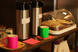 Auf einem Holztisch stehen zwei Thermoskannen, Tassen und ein Behälter mit Kuchen.
