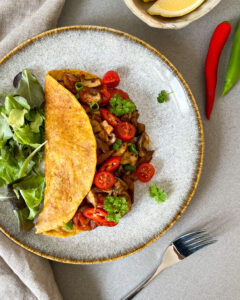 Auf einem Teller liegt das indische Omelett mit Salat, daruber eine kleine Schale mit einem Zitronenstück, darunter eine rote Chilischote und rechts unten im Bild liegt eine Gabel.