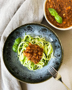 Auf einem blauen Teller befinden sich grüne "Zoodles" - also Spaghetti aus Zucchini. Oben drauf Soße Bolognese. Rechts daneben befindet sich in einer Schale der Rest der Bolognese. Ins Bild hinein ragt eine Gabel